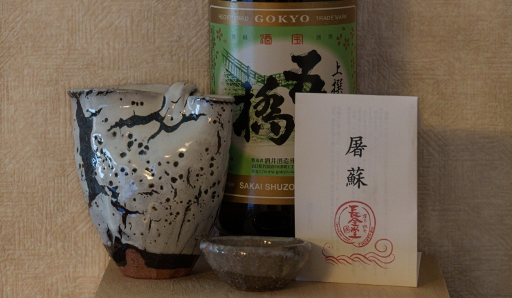 A ceramic sake pitcher and cup with a bottle of sake and a paper envelop of spices and herbs.