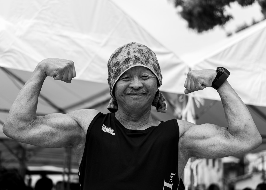 A smiling older man with a mustache. He is wearing a towel on his head, a large watch, and a black tanktop and is flexing his muscular arms.