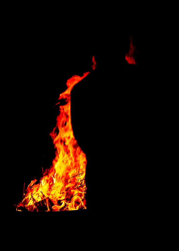 Everything is dark except a large fire burning high in the center. The silhouette of a person can just be made out in front of it.