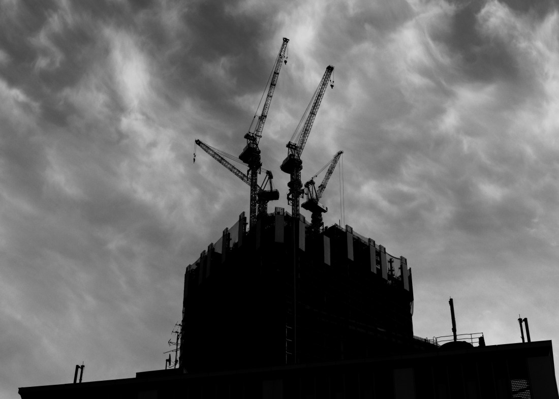 A black and white picture of gantry cranes atop a building under construction, against a sky streaked with whispy clouds