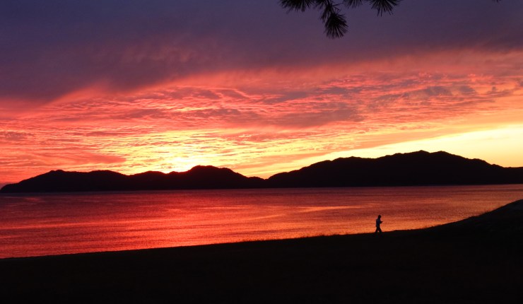 Sunset over the Seto Inland Sea. The sky is vivid orange fading to pink and purple. There is an island against the horizon. A figure in silhouette walks along the beach.