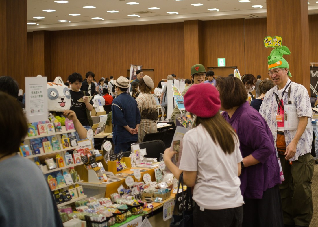 A crowd of people browsing tables lined with books. One man wears a bright green cap in the shape of some kind of plant character, and nearby a woman has a full-head papier mache cat mask on.