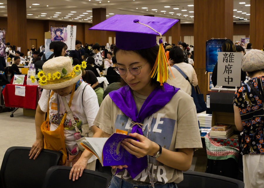 A Japanese woman with glasses in a t-shirt with a purple graduation cap is reading a purple book.