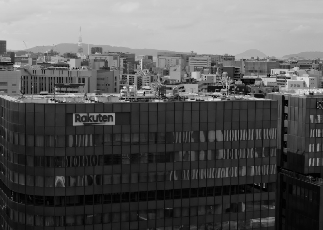 A monochrome shot of city rooftops. In the foreground is a building that is rectangular except the foreground left corner, which is rounded. It has a large "Rakuten" sign. A man in white shirt and black pants is walking acorss the roof.