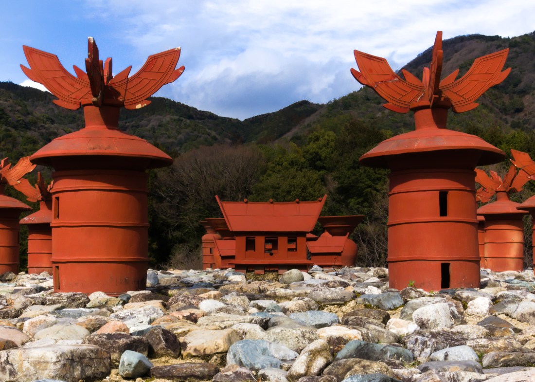 A low-angled shot of red ceramic buildinglike structures. Two towers (left and right) and a Japanese templelike building in the background. The perspective makes them look bigger than they are. Green hills are in the background.