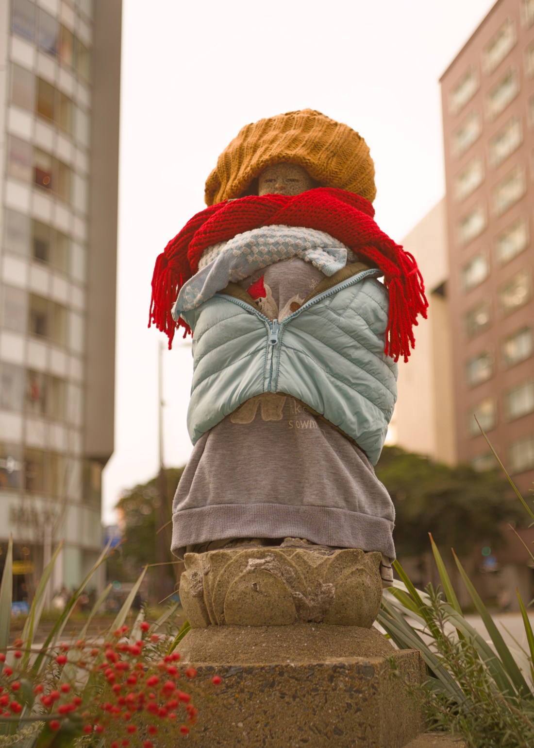 A small stone Jizo-sama statue on a city street corner. It is wrapped up in a child's coat, red knitted scarf, and pink knitted hat.